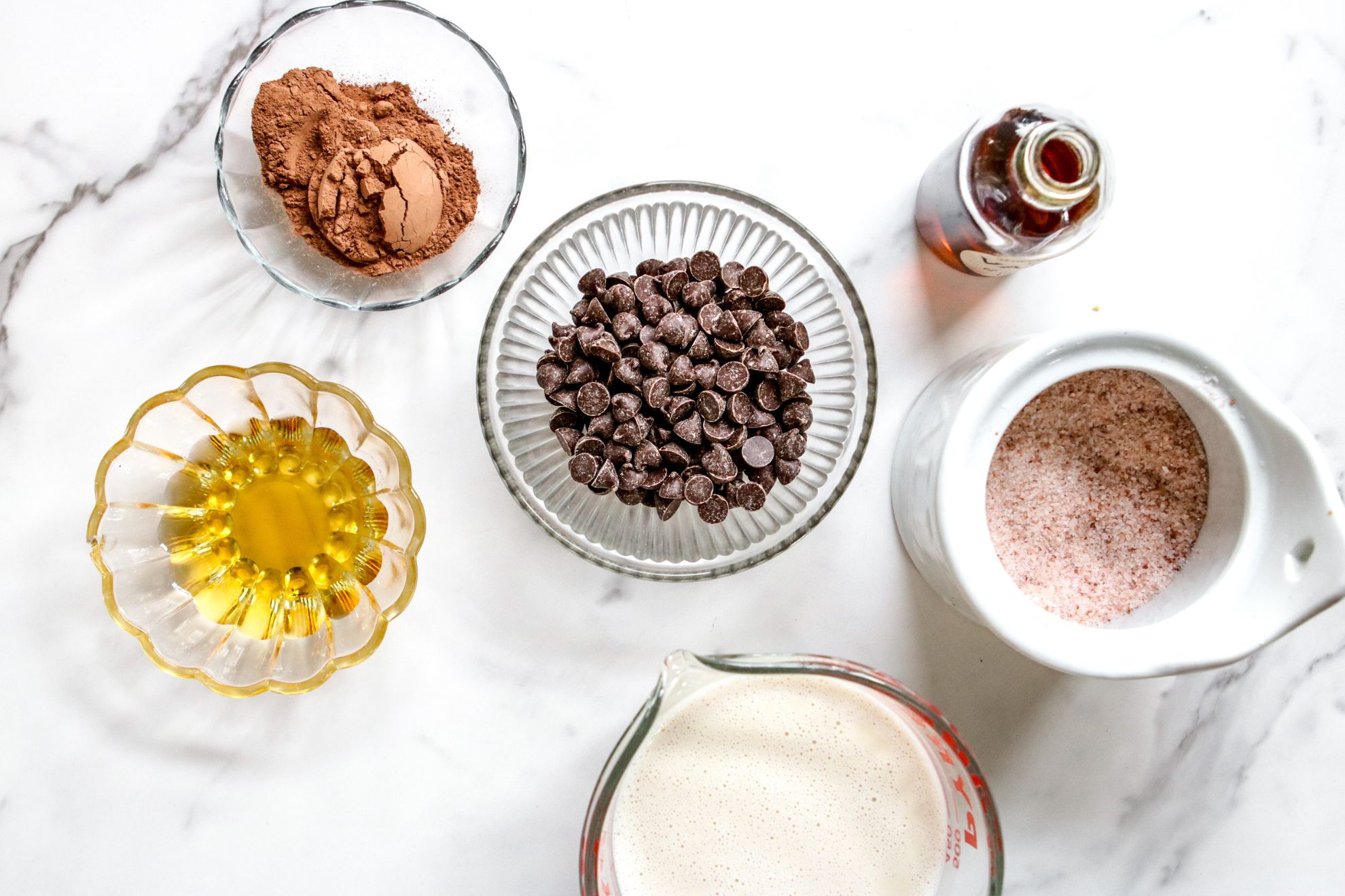 This is an overhead horizontal image of small bowls fitted with ingredients. There is a small bowl of cocoa powder, agave nectar, chocolate chips, a pyrex of oat milk, a small glass jar of vanilla, and a white ceramic container of pink salt.