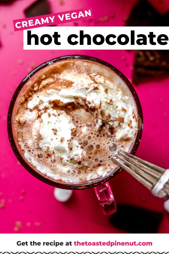 This is an overhead image of hot chocolate with whipped cream in a glass mug. A silver utensil is in the hot chocolate and leaning against the side of the mug. The mug sits on a hot pink surface with pieces of a broken chocolate bar on the pink surface surrounding the mug. Text overlay reads "creamy vegan hot chocolate get the recipe at thetoastedpinenut.com"
