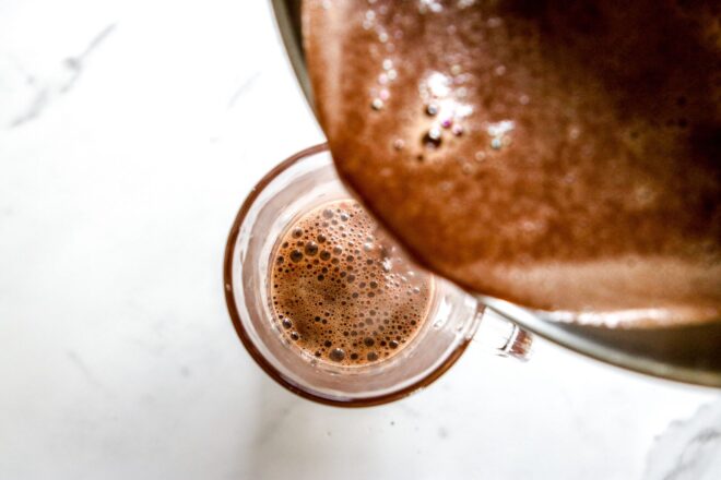 This is an overhead horizontal image of a glass mug sitting on a white marble surface. A saucepan with hot chocolate is overhead the being poured into the mug.