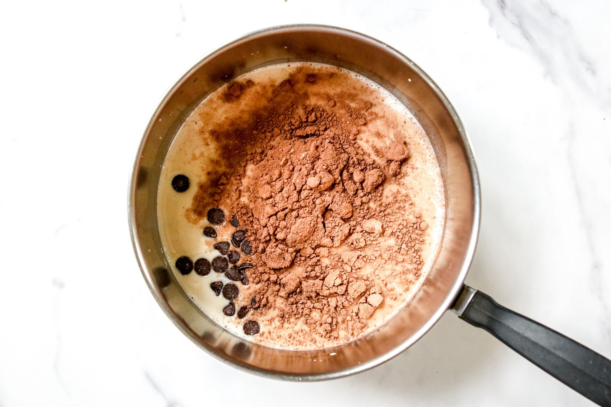 This is an overhead horizontal image of a saucepan sitting on a white marble surface. In the saucepan is oat milk, cocoa powder, and chocolate chips.