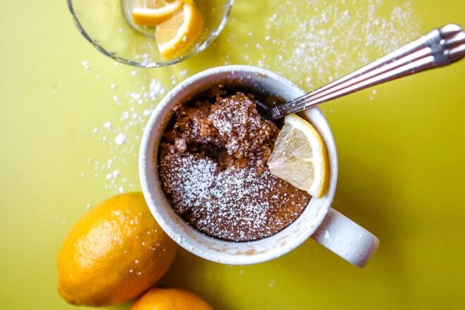 This is an overhead horizontal image of a white mug with a cooked mug cake sprinkled with powdered sugar. A lemon wedge is on top of the mug and a silver spoon is digging into the cake. The mug sits on a yellow surface with powdered sugar sprinkled around it. Two lemons are next to the mug to the bottom left of the image. A small glass bowl with more lemon wedges is to the top left corner of the image.