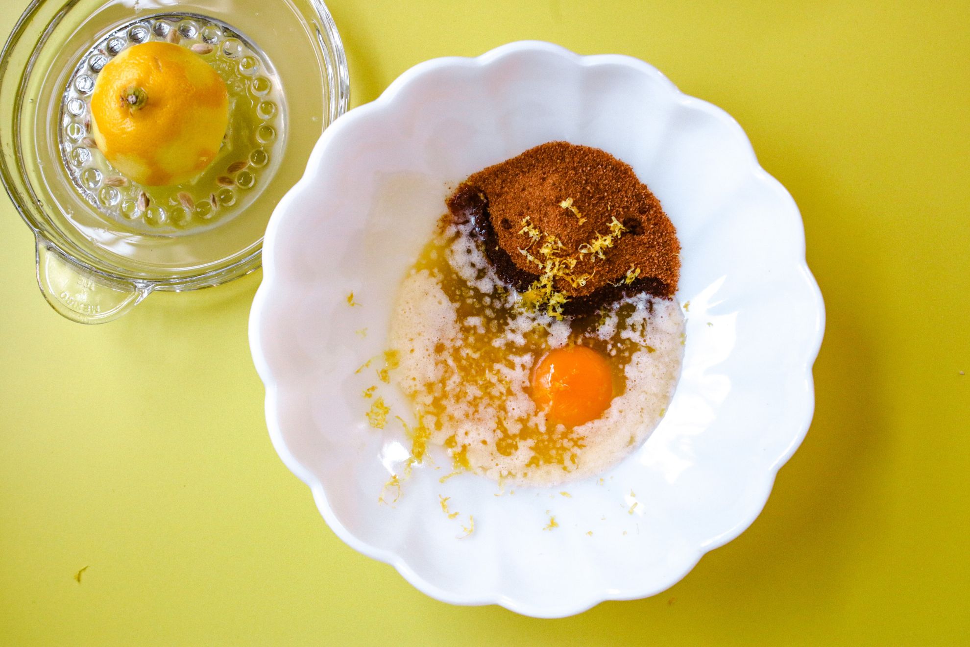 This is an overhead horizontal image of a white scalloped bowl with ingredients in it like coconut sugar, lemon zest, butter, and an egg. The bowl sits on a yellow surface with a glass juicer to the top left corner. The juicer has half of a lemon on it.