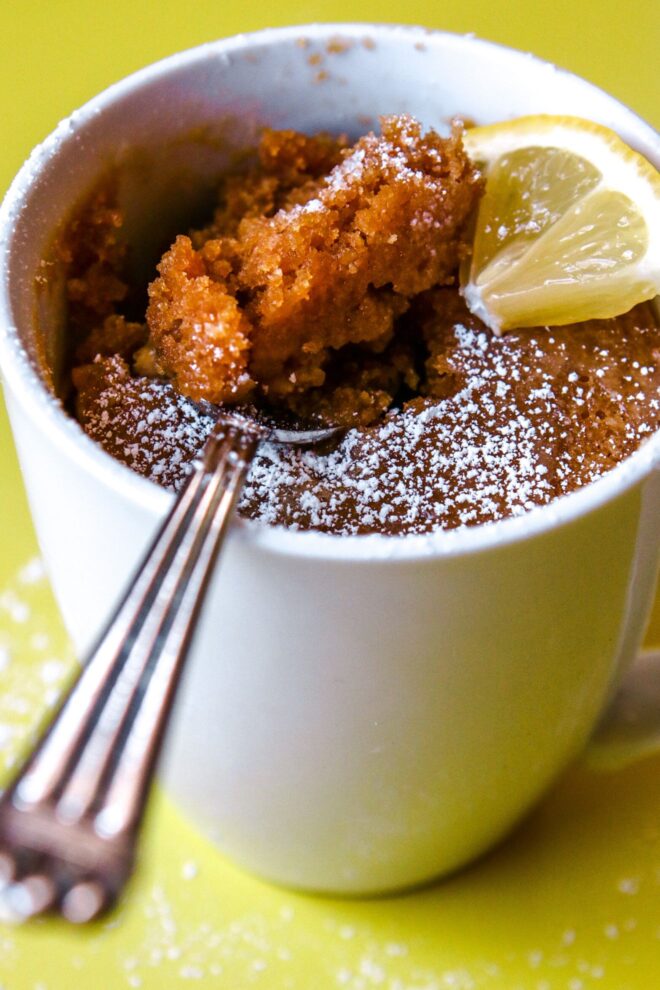 This is vertical image looking at a white mug with baked cake in it from a side view. The cake is sprinkled with powdered sugar and a lemon wedge is to the right side of the mug. A silver spoon is digging into the cake. The mug sits on a yellow surface with more powdered sugar sprinkled around.