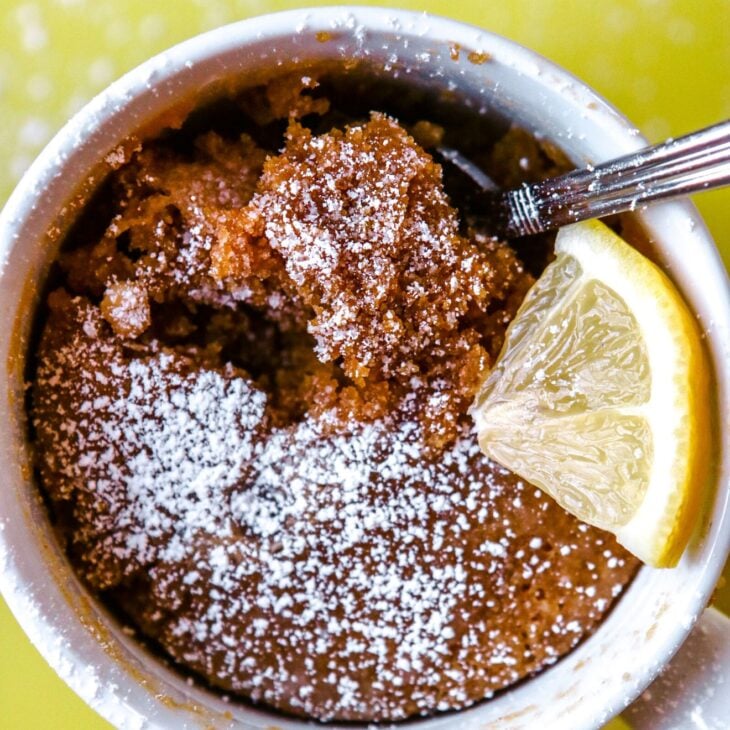 This is an overhead vertical image of a white mug with baked cake in it. The cake is sprinkled with powdered sugar and a lemon wedge is to the right side of the mug. A silver spoon is digging into the cake. The mug sits on a yellow surface with more powdered sugar sprinkled around.