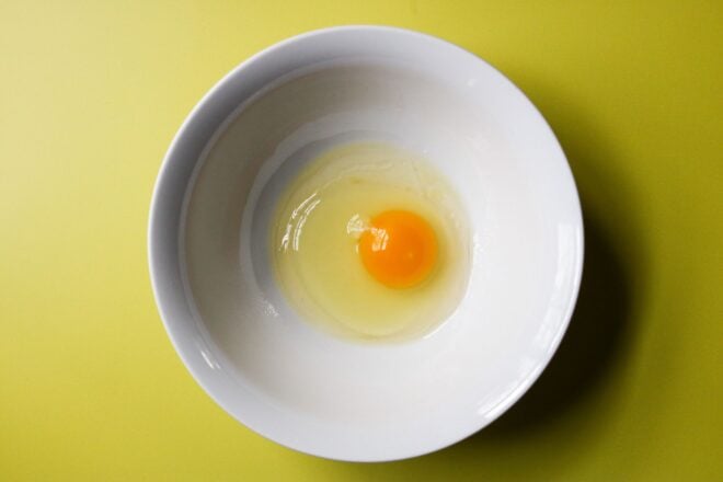 This is an overhead horizontal image of a white bowl with a raw egg in the center. The bowl sits on a yellow surface.