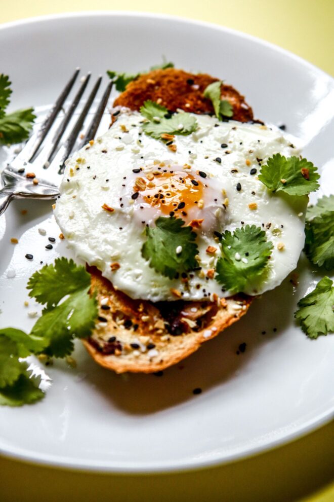 This is a vertical image of a fried egg on a slice of toast. The egg is topped with cilantro and everything bagel seasoning. The toast is on a white plate with more cilantro leaves surrounding the toast and a silver fork leaning against the side to the left of the image. The plate sits on a bright yellow surface.