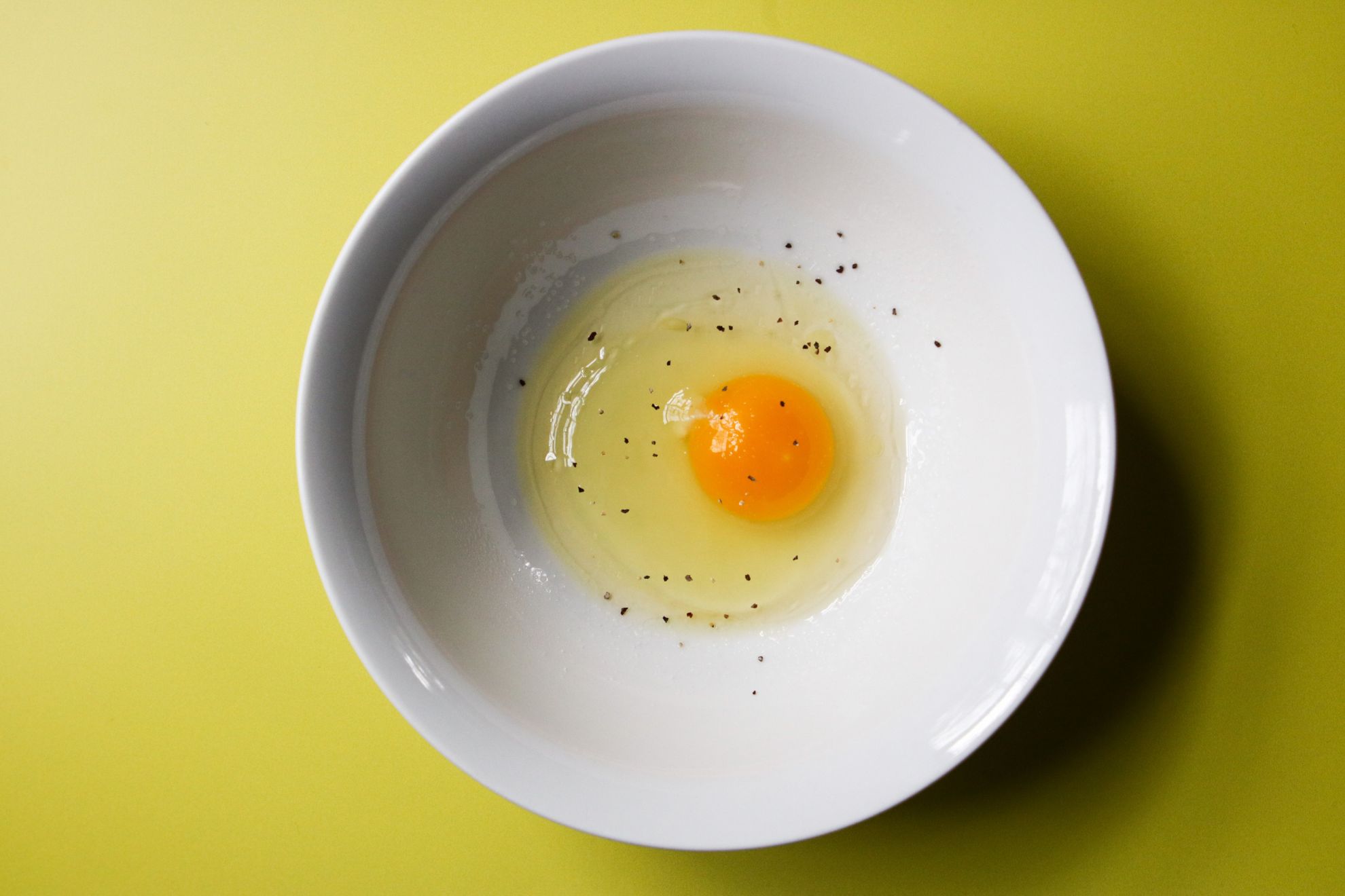 This is an overhead horizontal image of a white bowl with a raw egg in the center topped with pepper. The bowl sits on a yellow surface.