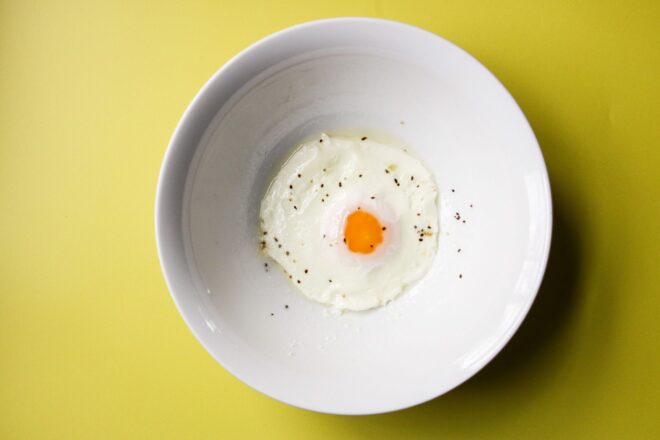 This is an overhead horizontal image of a white bowl with a fried egg in the center topped with pepper. The bowl sits on a yellow surface.