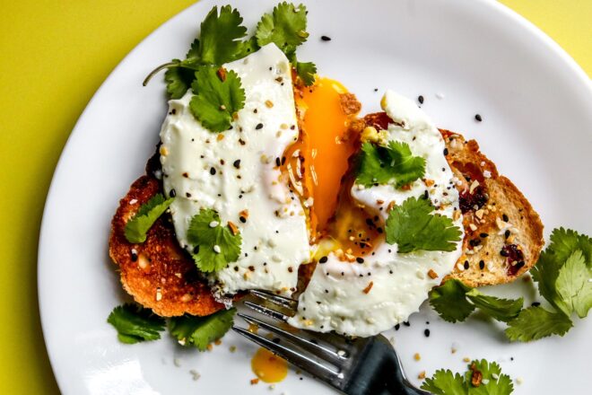 This is an overhead horizontal image of a drippy fried egg on a slice of toast. The egg is topped with cilantro and everything bagel seasoning. The toast is on a white plate with more cilantro leaves surrounding the toast and a silver fork leaning against the side to the bottom center of the image. The toast and egg is cut in half with a drippy yellow yolk on the plate. The plate sits on a bright yellow surface.
