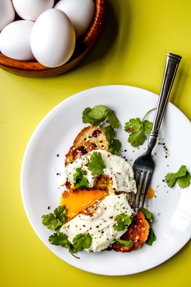 This is an overhead vertical image of a drippy fried egg on a slice of toast. The egg is topped with cilantro and everything bagel seasoning. The toast is on a white plate with more cilantro leaves surrounding the toast and a silver fork leaning against the side. The toast and egg is cut in half with a drippy yellow yolk on the plate. The plate sits on a bright yellow surface with a wood bowl of whole white eggs in it.