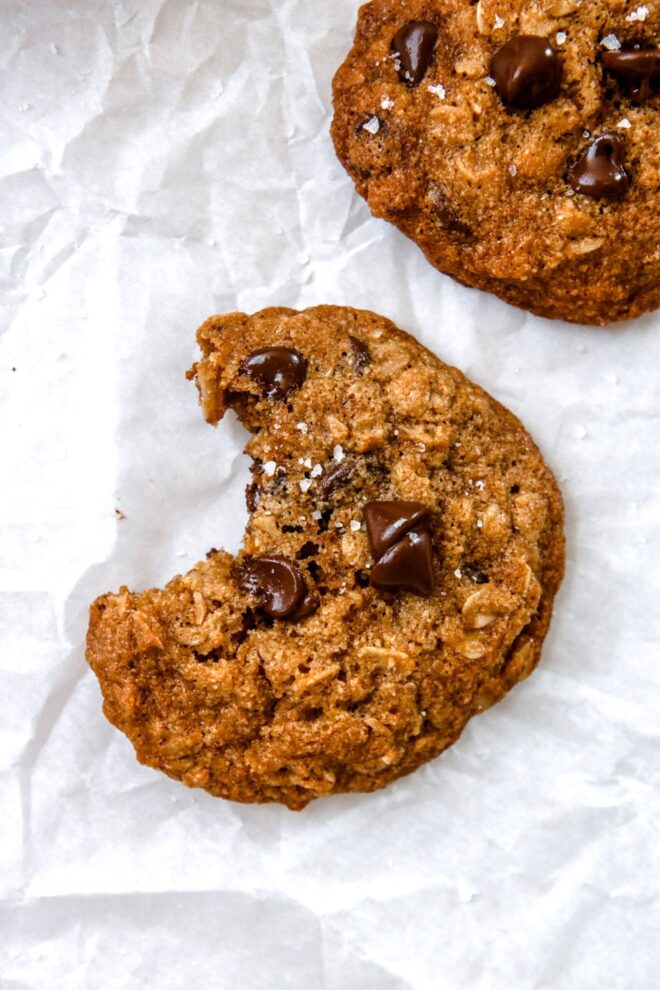 This is an overhead vertical image of an oatmeal chocolate chip cookie with a bite taken out of it. The cookie is sprinkled with flakey salt and sits on a white piece of parchment paper. Another cookie is in the top right corner of the image.
