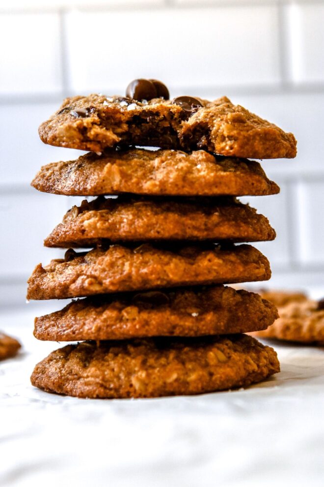 This is a vertical image of a stack of six chocolate chip cookies. The image shows the stack from the side and the top cookie has a bite taken out of it. The stack sits on a light surface with more cookies blurred in the background.