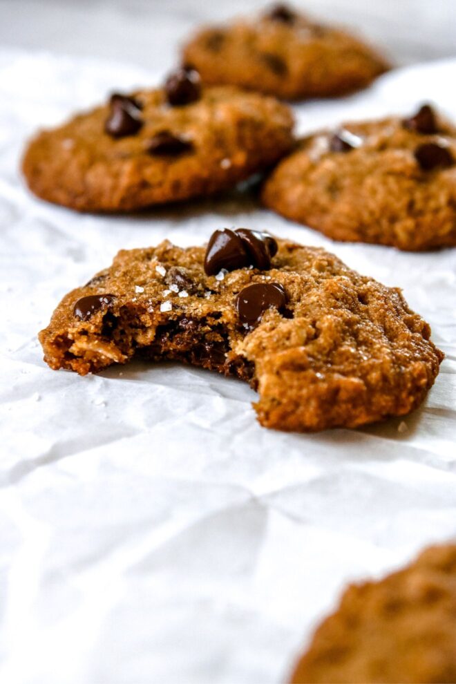 This is a vertical image of baked oatmeal chocolate chip cookies. The cookies are topped with flakey salt and sit on a crumpled piece of white parchment paper. This is a side view focusing on one cookie with a bite taken out of it. The more cookies are blurred in the background behind the main bitten cookie.