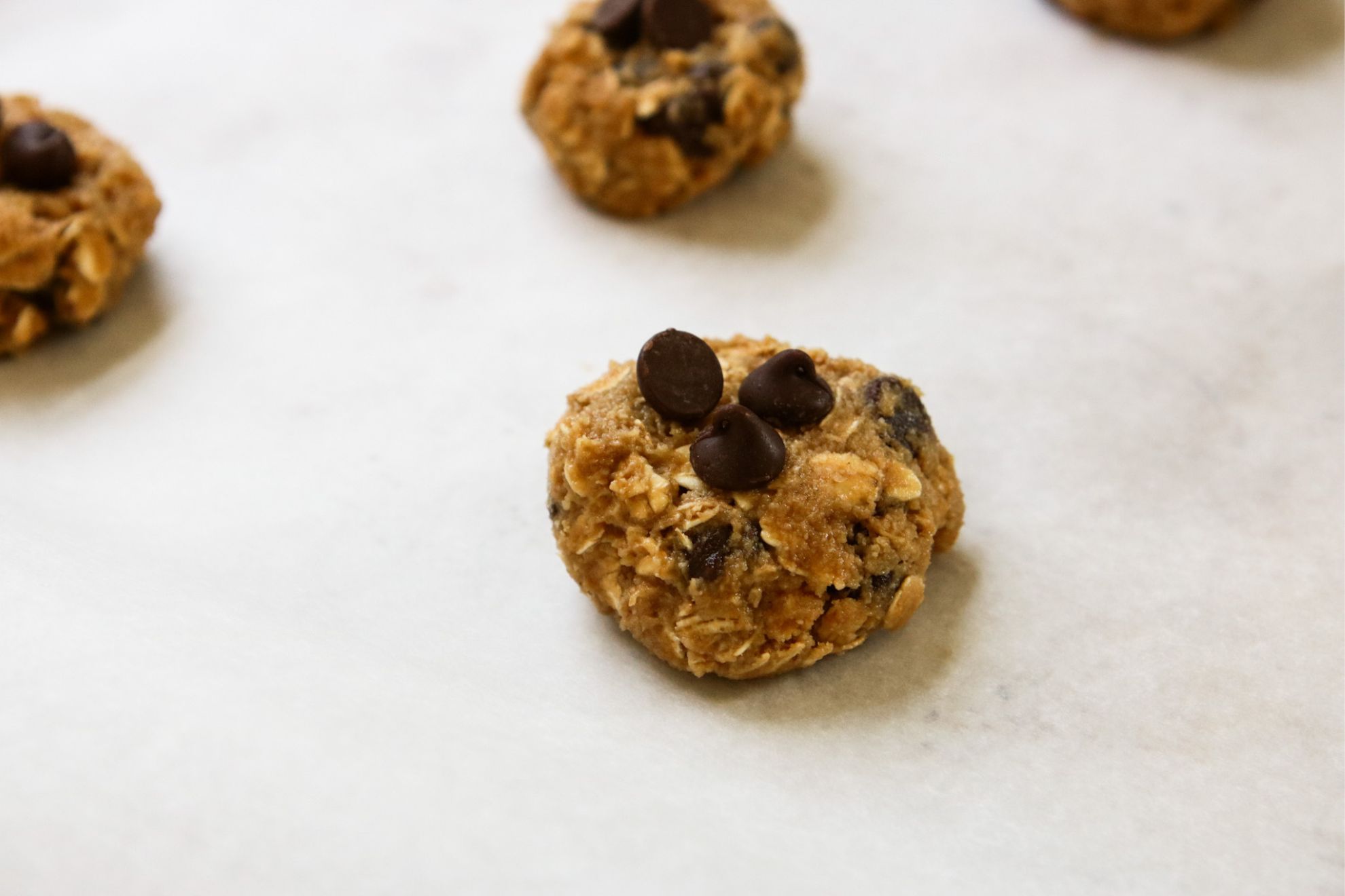 This is a horizontal image showing the side view of a flattened oatmeal cookie dough ball topped with chocolate chips. The cookie dough is raw and sits on a piece of white parchment paper. More cookie dough are in the background behind the main one in focus.