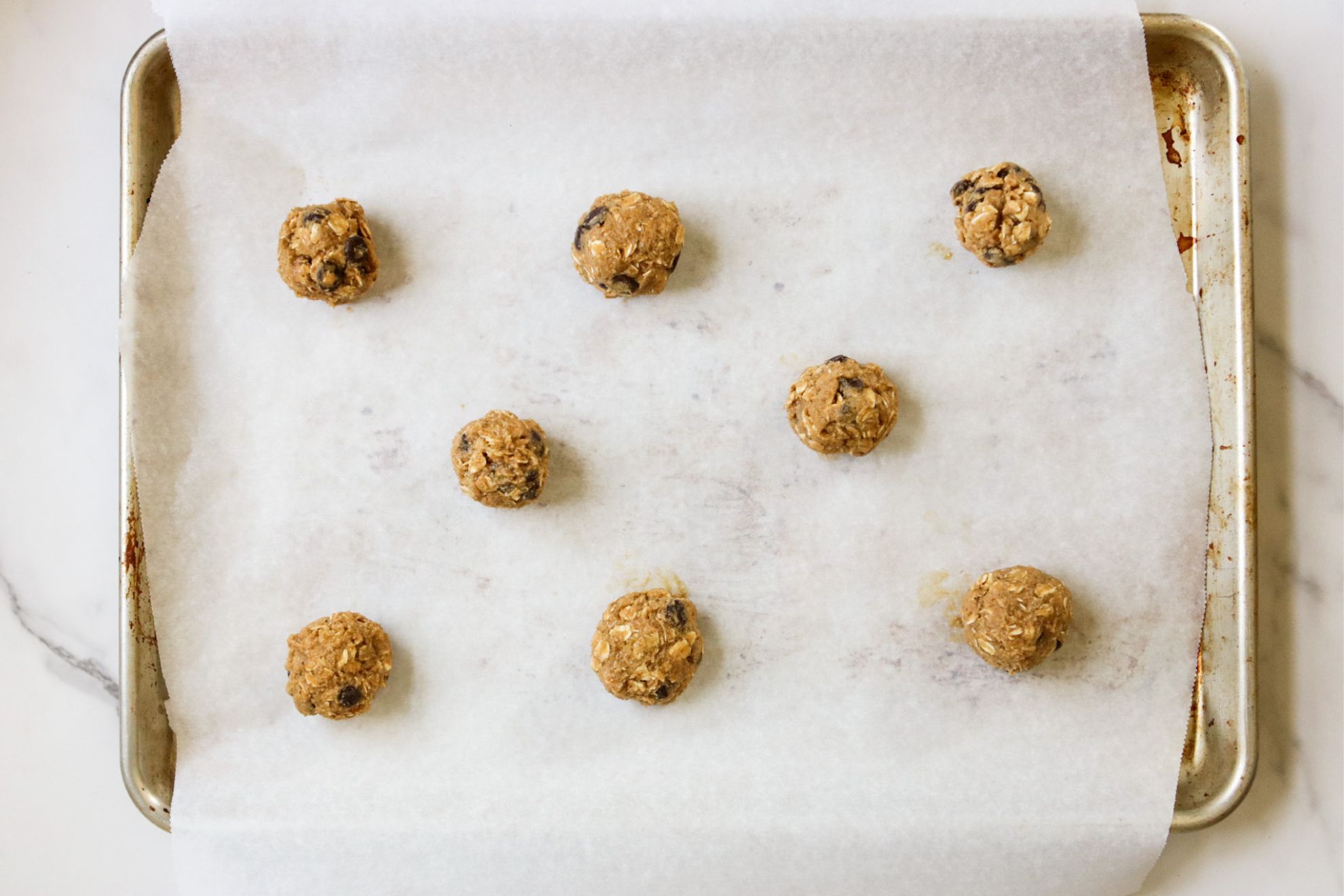 This is an overhead horizontal image of a baking sheet lined with parchment paper and eight cookie dough balls spaced a few inches apart. The pan sits on a light marble surface.