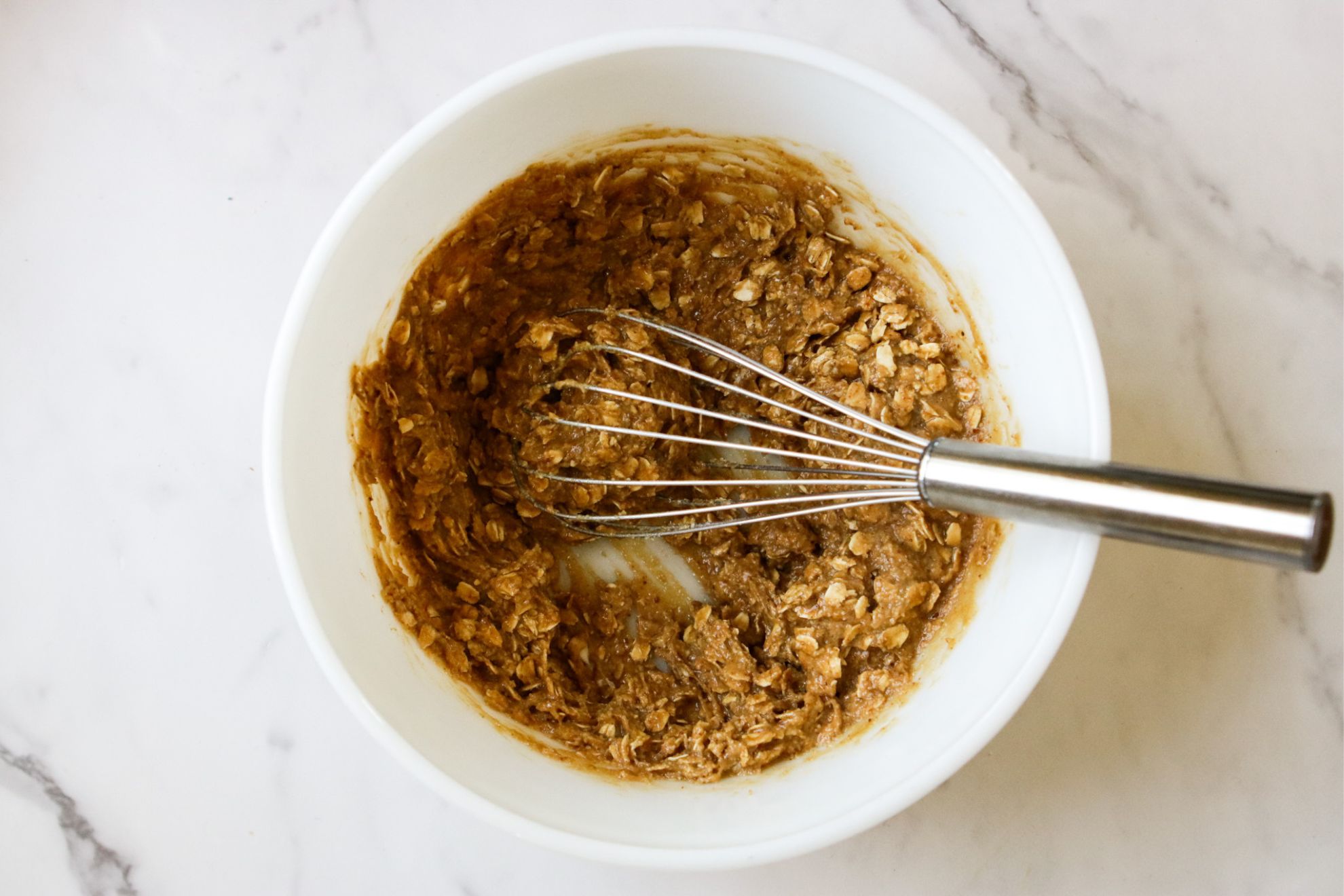 This is an overhead horizontal image of a white bowl on a white marble surface. In the bowl is an oatmeal cookie dough mixed together. A whisk is inserting into the ingredients and leaning against the side of the bowl with the whisk pointing to the right side of the image.