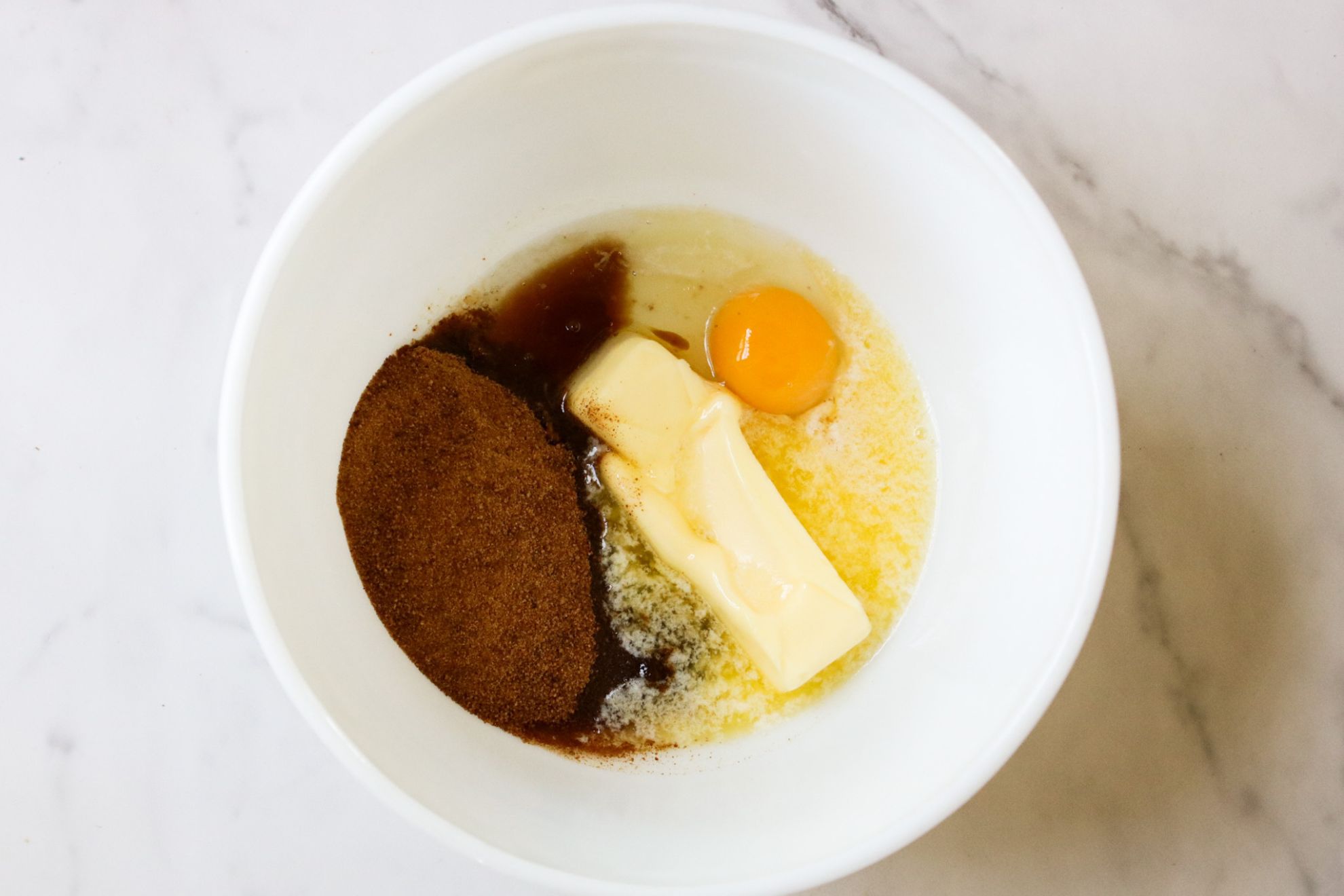 This is an overhead horizontal image of a large white bowl on a white marble surface. The bowl has softened butter, an egg, coconut sugar, and vanilla extract in it.