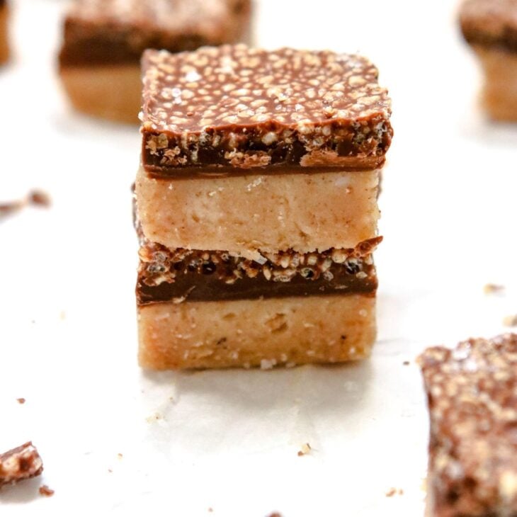 This is a vertical image looking at a 3/4 view onto two banana bites stacked ontop of each other. The banana bites have two layers. The bottom layer is a light beige color and the top layer is a hardened chocolate with crispy quinoa. More banana bites are in front and back of the stacked two.