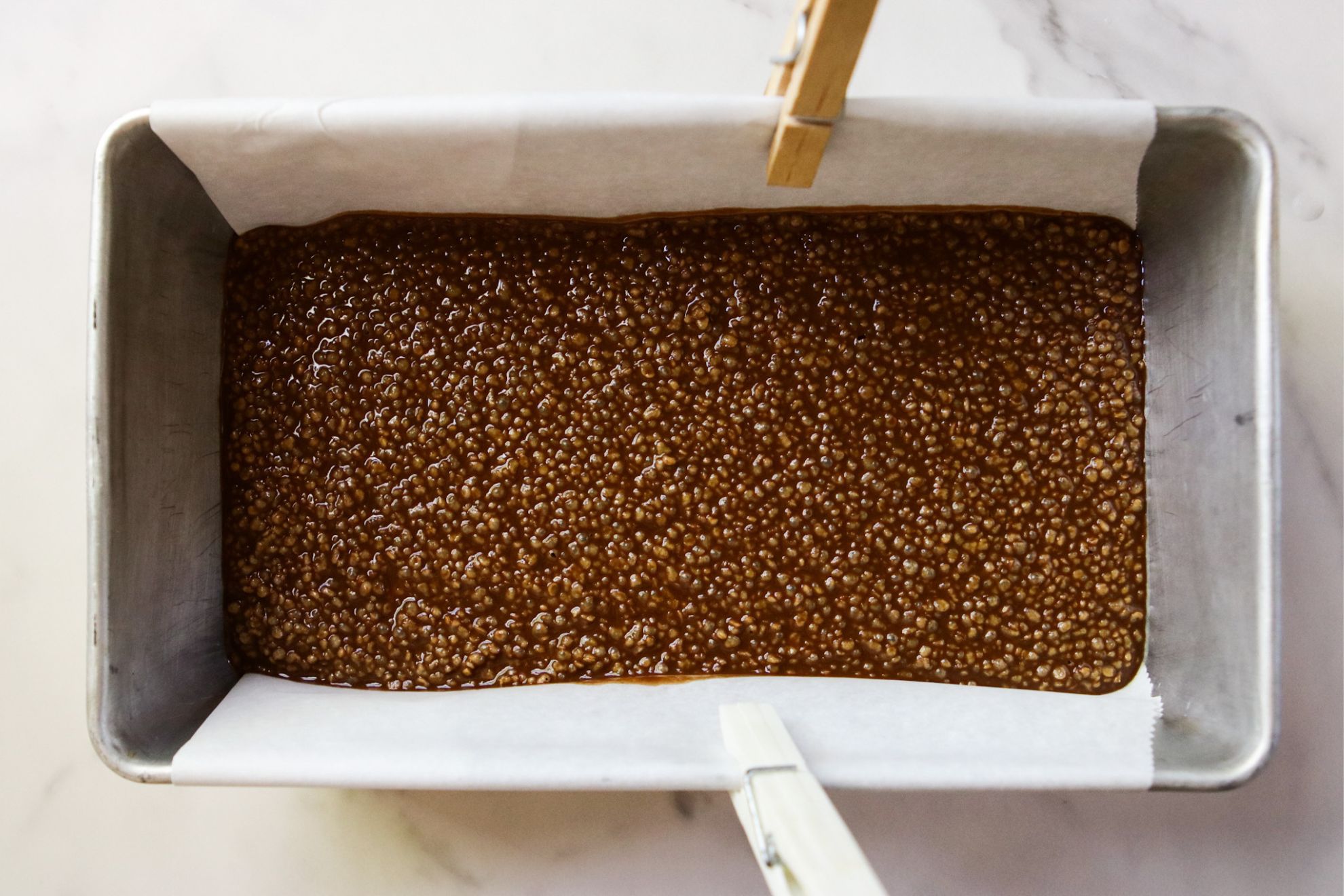 This is an overhead horizontal image of a bread pan lined with parchment paper hanging over the long two sides of the pan. The parchment paper is secured by clothespins on either side. In the pan is a chocolate puffed quinoa mixture spread across the bottom.