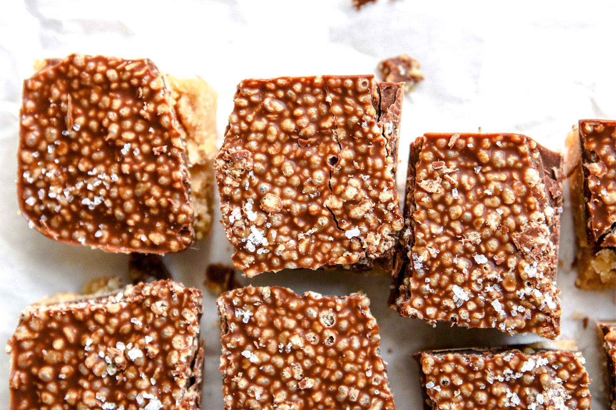 This is an overhead horizontal image of small squares on a white piece of parchment paper. The squares are a harden chocolate with crispy quinoa.