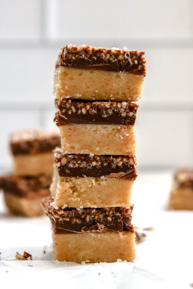 This is a side view of a stack of four banana bites on a piece of parchment paper with more blurred in the background. The banana bites have two layers to them, a bottom beige color and the top is a hardened chocolate with crispy quinoa.
