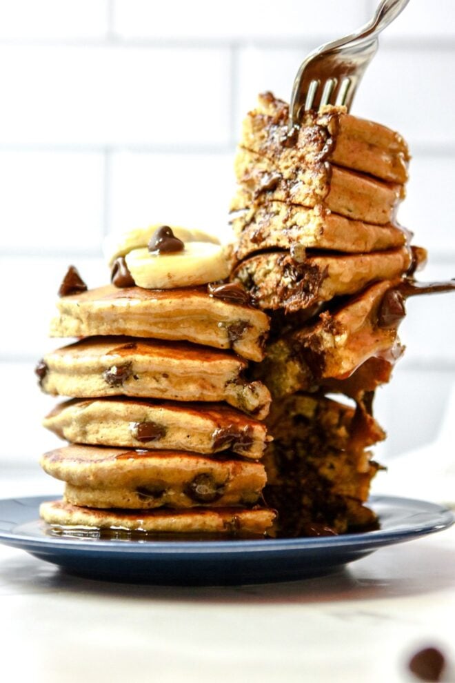 This is a vertical image of a side view of a stack of chocolate chip pancakes. The stack sits on a dark blue plate. The plate sits on a white surface with white subway tile in the background. The stack of pancakes is topped with sliced banana and chocolate chips. A fork is coming in from the top of the image and lifting up a wedge of cut pancakes from the stack.