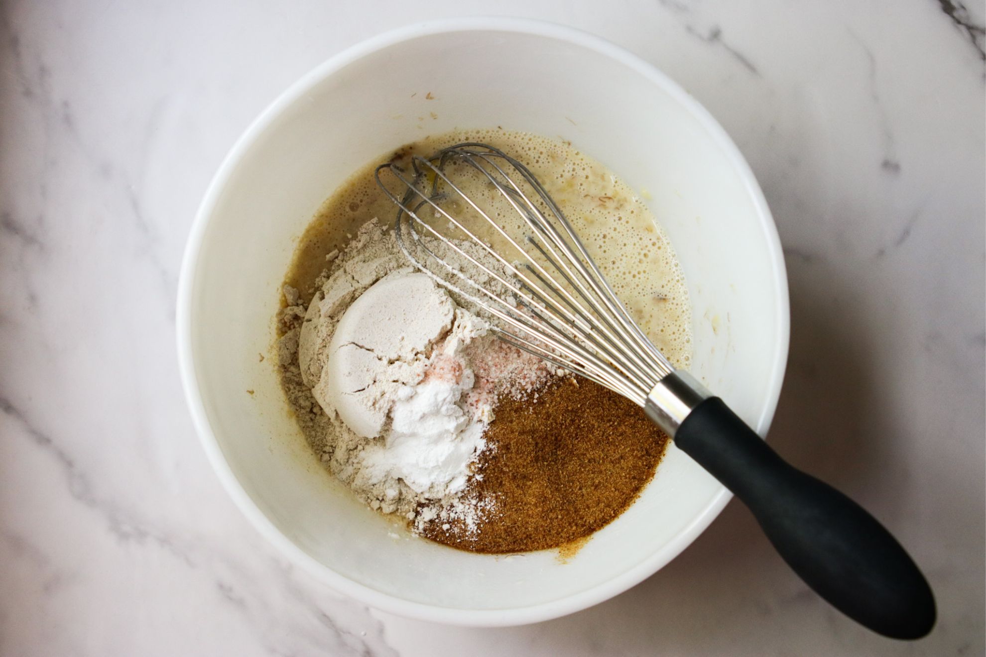 This is an overhead horizontal image of wet ingredients with dry ingredients on top. The ingredients are in a white bowl on a marble counter. A whisk with a black handle is in the bowl with the black handle leaning against the side pointing to the bottom right of the image.