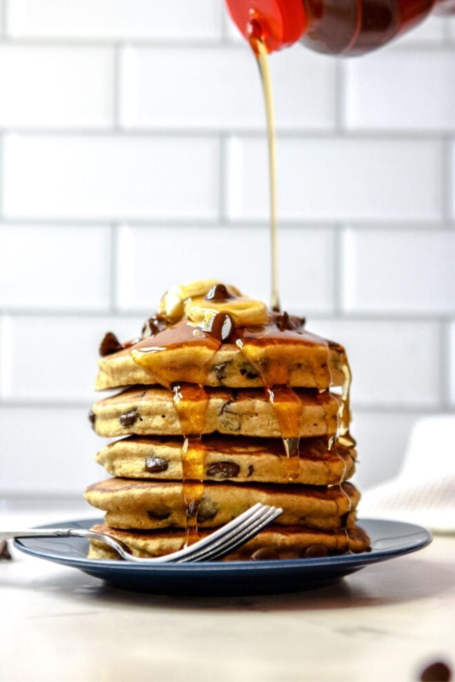 This is a vertical image of a side view of a stack of chocolate chip pancakes. The stack sits on a dark blue plate with a fork at the bottom of the stack leaning against the side of the plate. The plate sits on a white surface with white subway tile in the background. The stack of pancakes is topped with sliced banana and chocolate chips. A syrup bottle with a red lid is coming from the top of the image and pouring syrup on top of the pancakes. The syrup drips down the sides of the pancake stack.