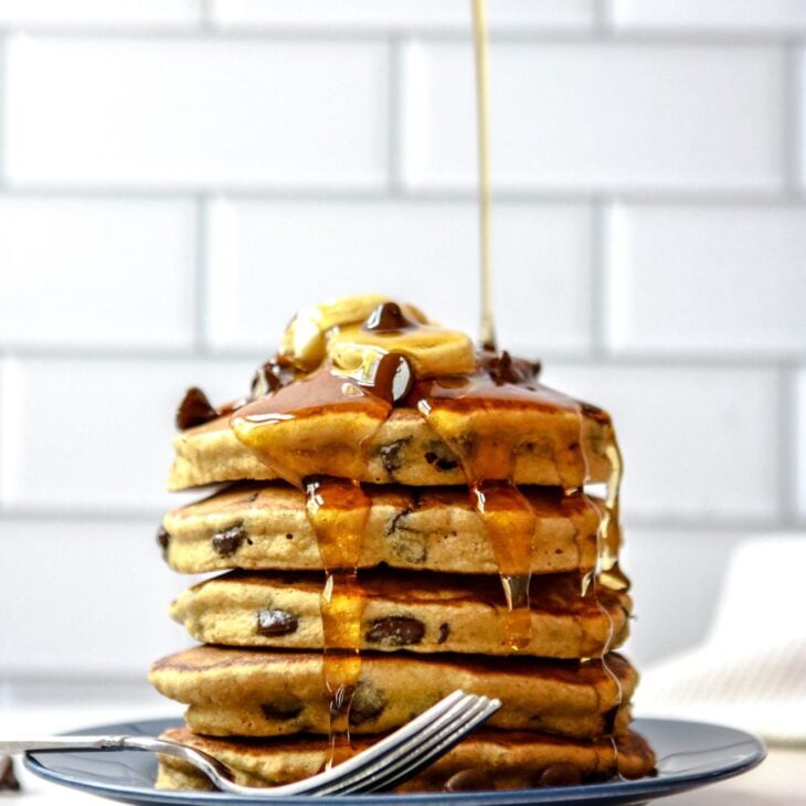 This is a vertical image of a side view of a stack of chocolate chip pancakes. The stack sits on a dark blue plate with a fork at the bottom of the stack leaning against the side of the plate. The plate sits on a white surface with white subway tile in the background. The stack of pancakes is topped with sliced banana and chocolate chips. A syrup bottle with a red lid is coming from the top of the image and pouring syrup on top of the pancakes. The syrup drips down the sides of the pancake stack.