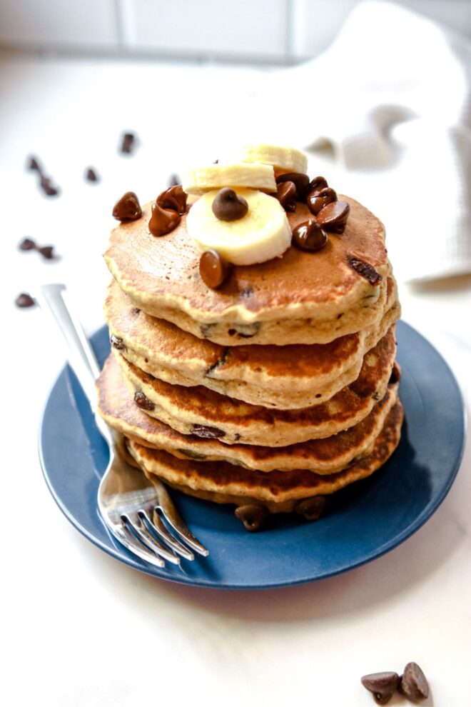 This is a vertical image of a 3/4 view of a stack of chocolate chip pancakes. The stack sits on a dark blue plate with a fork at the bottom of the stack leaning against the side of the plate. The plate sits on a white surface with white subway tile in the background. The stack of pancakes is topped with sliced banana and chocolate chips.