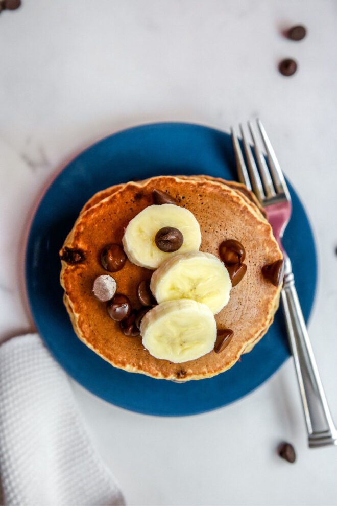This is an overhead vertical image looking down on a stack of pancakes on a blue plate. The pancakes are topped with sliced banana and chocolate chips. A fork is on the right side of the plate. The plate sits on a white marble surface with a couple chocolate chips scattered around and a knitted cloth napkin to the bottom left of the image.
