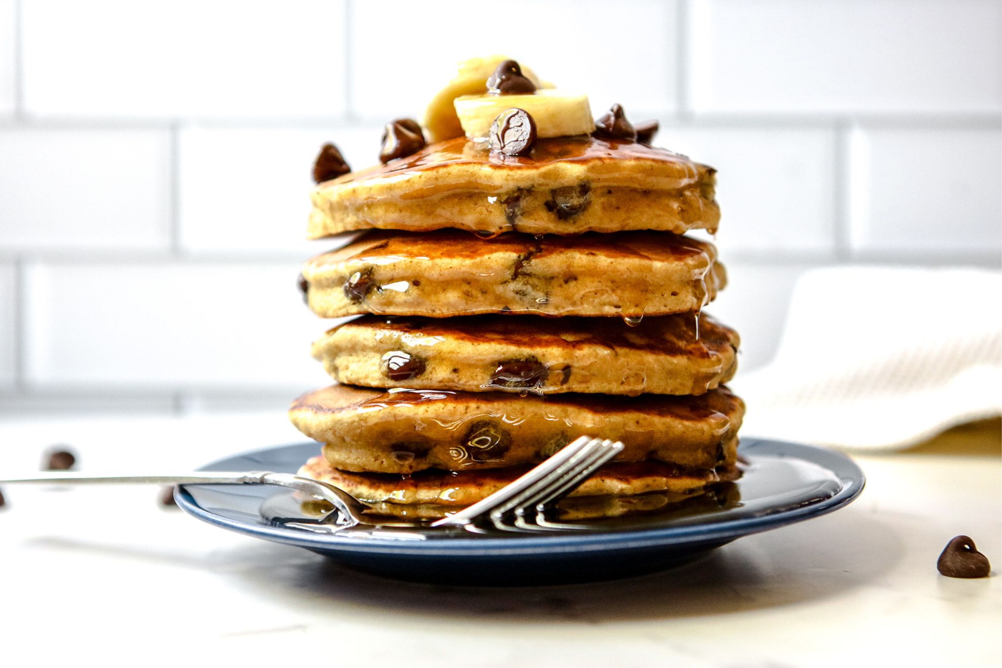 This is a horizontal image of a side view of a stack of chocolate chip pancakes. The stack sits on a dark blue plate with a fork at the bottom of the stack leaning against the side of the plate. The plate sits on a white surface with white subway tile in the background. The stack of pancakes is topped with sliced banana and chocolate chips.