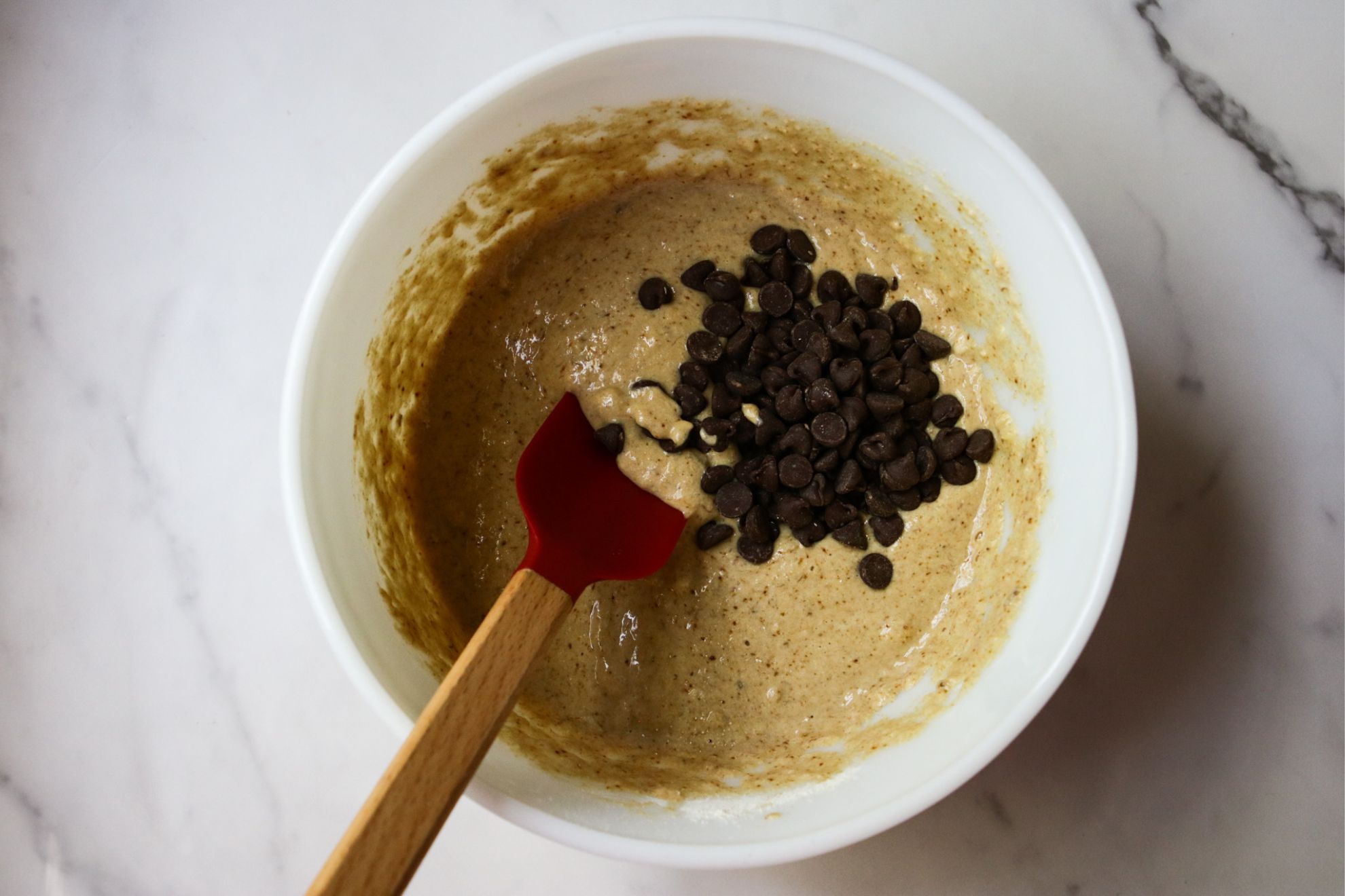 This is an overhead horizontal image of mixed batter with chocolate chips in a mound to the right side of the bowl. The bowl is white and sits on a white marble counter. A red spatula with a wooden handle is dipping into the batter and leaning against the side of the bowl, pointing to the bottom left of the image.