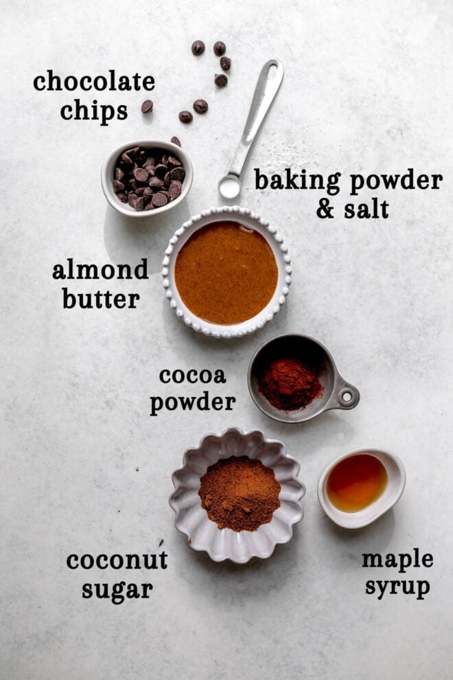 This is an overhead image of ingredients in small individual bowls on a light grey surface. Each ingredient has text overlay next to it that labels it: "chocolate chips, baking powder & salt, almond butter, cocoa powder, coconut sugar, and maple syrup."
