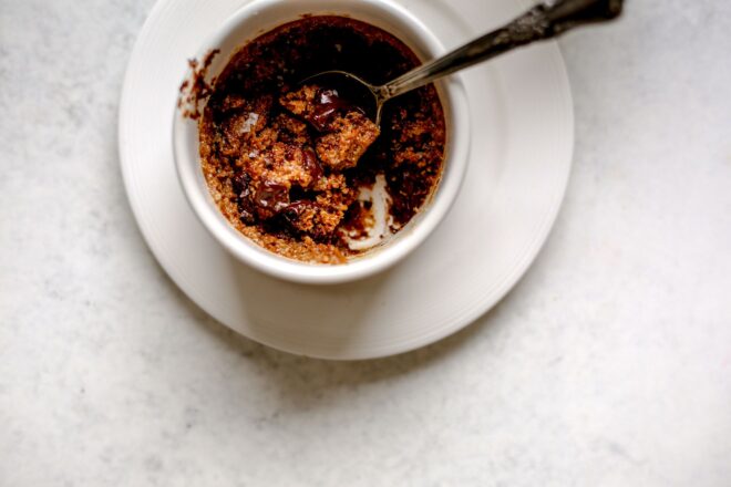 This is an overhead horizontal image of a white ramekin on a white plate sitting on a light grey surface. In the ramekin is a chocolate chip cookie with an antique spoon dipping into it and leaning against the right side of the ramekin.