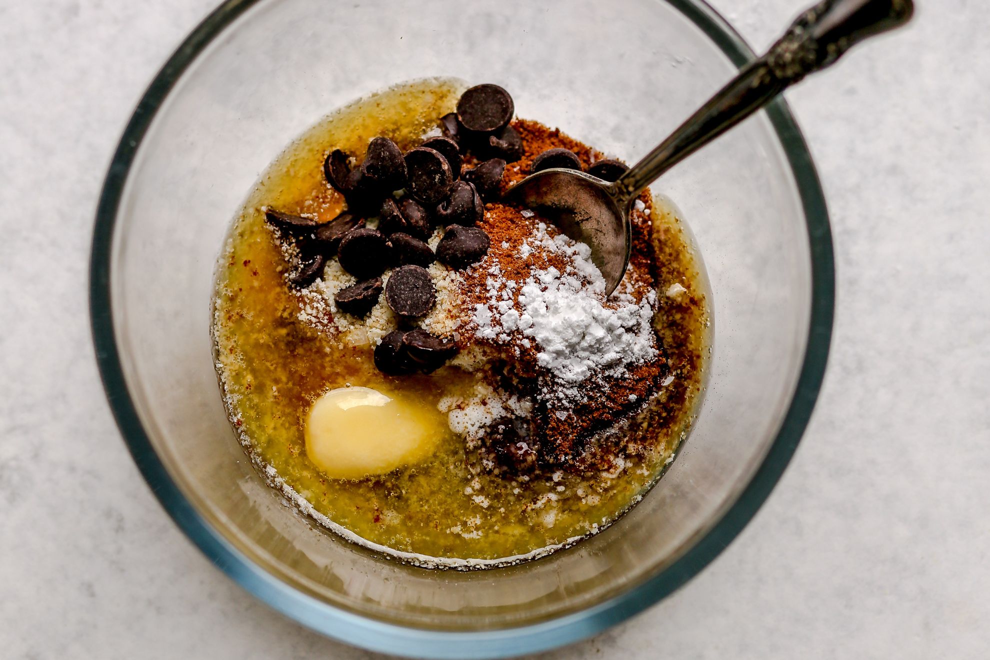 This is an overhead horizontal image of ingredients like melted butter, chocolate chips, coconut sugar, and baking powder in a glass bowl. A spoon is in the bowl, leaning against the right side. The bowl sits on a light grey surface.