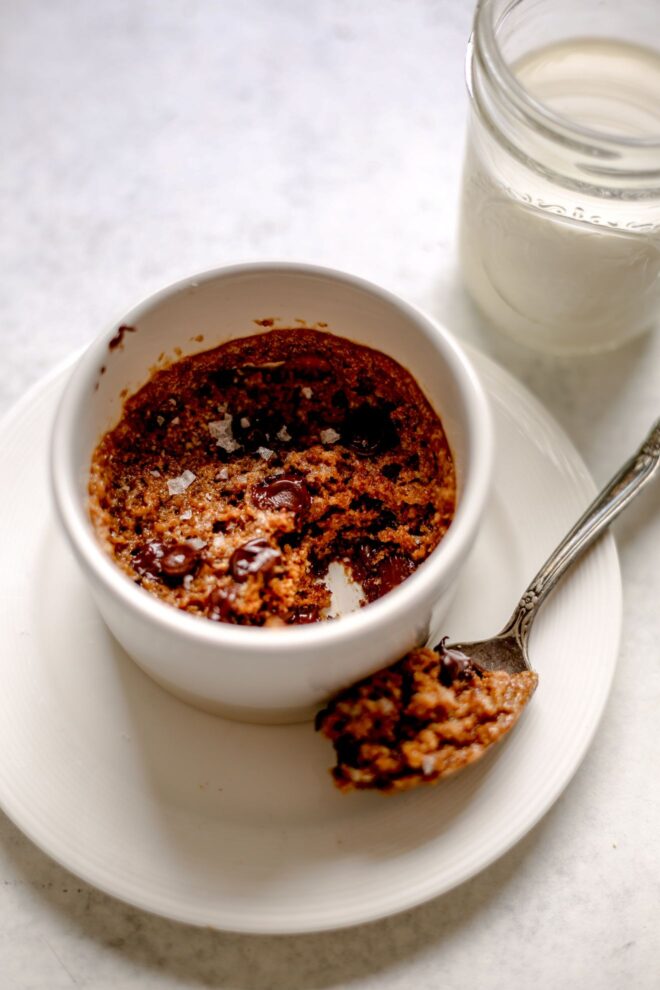This is an overhead vertical image of a white ramekin on a white plate sitting on a light grey surface. In the ramekin is a chocolate chip cookie with an antique spoon with a big scoop of cookie leaning on the plate next to the ramekin. A small mason jar of milk is in the top right corner of the image.