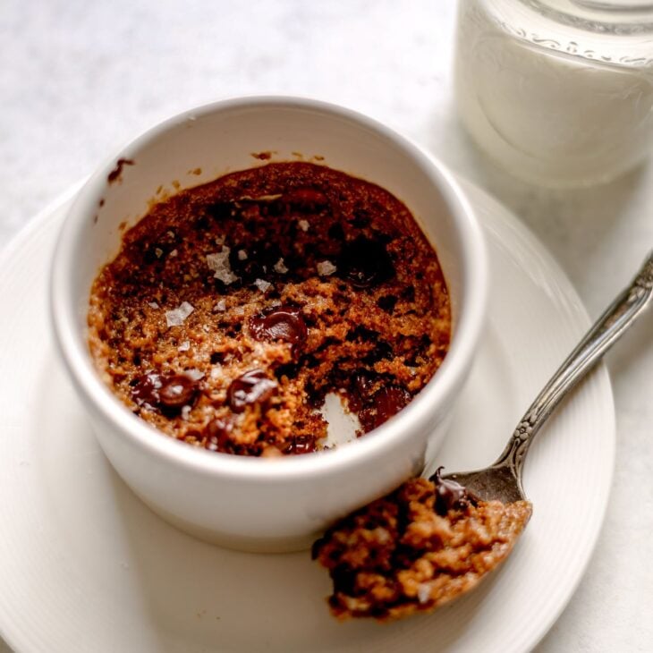 This is an overhead vertical image of a white ramekin on a white plate sitting on a light grey surface. In the ramekin is a chocolate chip cookie with an antique spoon with a big scoop of cookie leaning on the plate next to the ramekin. A small mason jar of milk is in the top right corner of the image.