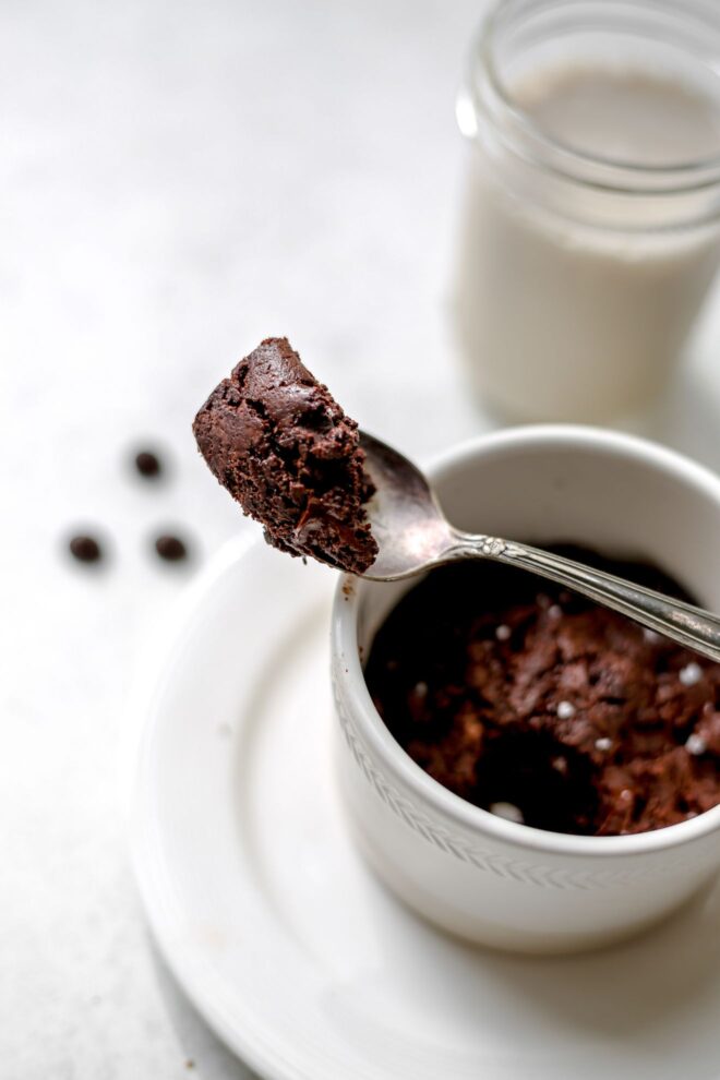 This is a vertical image looking at a side view of a silver spoon with fudgey chocolate brownie leaning against a white mug. The white mug is on a white plate and some chocolate chips and a glass of milk are blurred in the background.