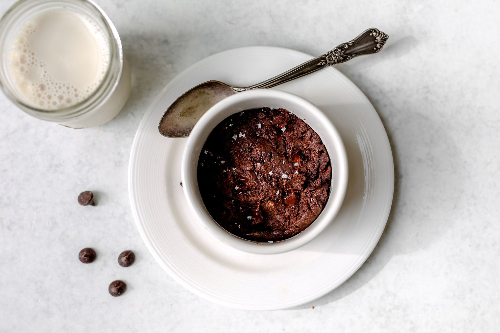 This is a horizontal overhead shot of a white ramekin with a brownie in it. The ramekin sits on a white plate with a silver spoon leaning against the side. The plate sits on a light grey surface with four chocolate chips to the left of it and a glass of milk to the top left corner of the image.