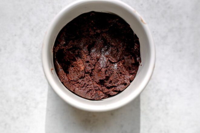 This is an overhead horizontal image of a white ramekin with raw brownie batter in it. The ramekin sits on a light grey surface and a soft shadow is projecting from the bottom of the ramekin to the bottom of the image.