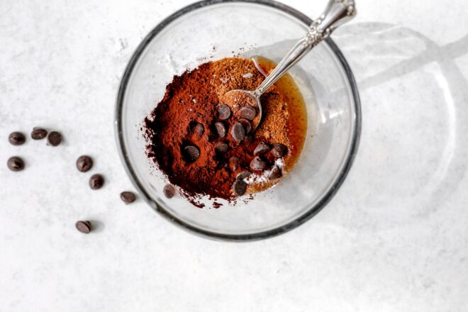 This is an overhead horizontal image of a glass bowl with ingredients in it. the ingredients appear to be cocoa powder, coconut sugar, and some chocolate chips. A silver spoon is dipping into the ingredients and leaning against the top right side of the bowl. The bowl is centered but towards the top of the image. Additional chocolate chips are scattered to the left of the bowl on the light grey surface.