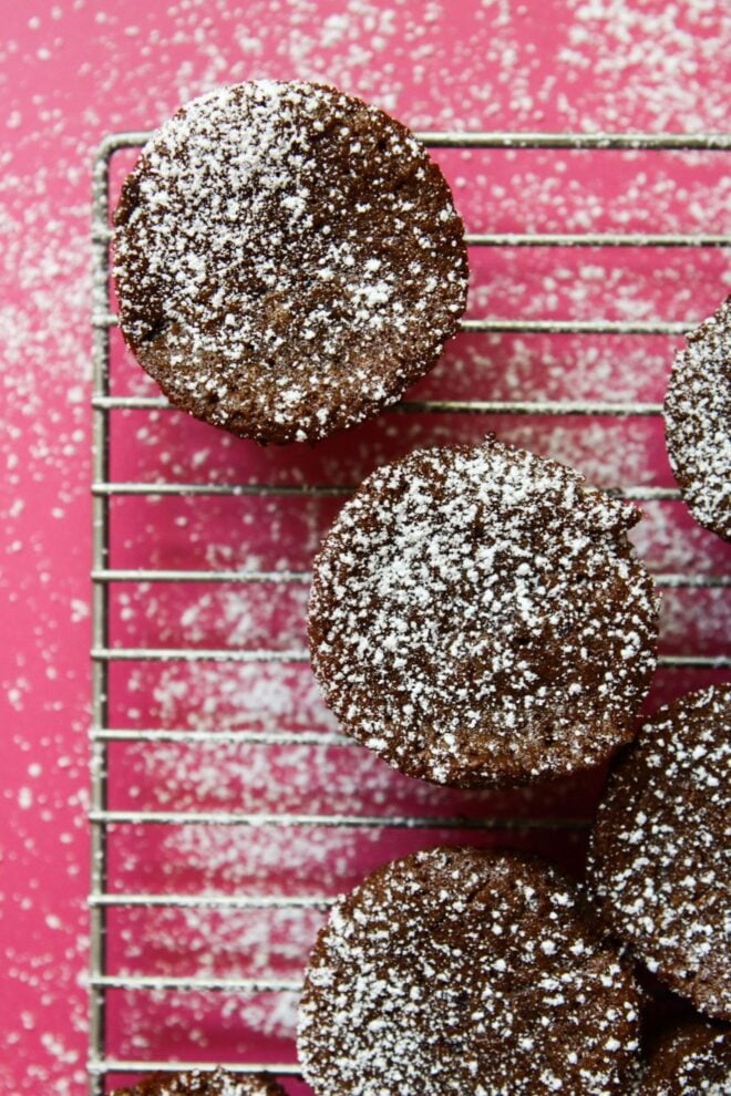 This is an overhead vertical image of circle shaped brownies on a cooling rack. The cooling rack is on a dark pink surface and the brownies are sprinkled with powdered sugar.
