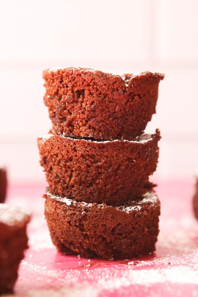 This is a vertical image shot from the side of a stack of three circular brownie bites. The stack is sitting on a pink surface and are dusted with powdered sugar. More bites are blurred around the stack and powdered sugar is dusting the pink surface. Behind the stack is a blurred white tile background.