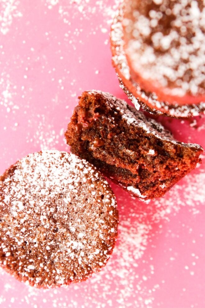 This is an overhead vertical image looking down onto small circle shaped brownies. The brownies are on a pink surface and dusted with powdered sugar. One brownie is in the left corner of the image and there is a stack of brownies to the top right corner. In the middle is a brownie with a bite taken out turned on its side to reveal its fudgey center.