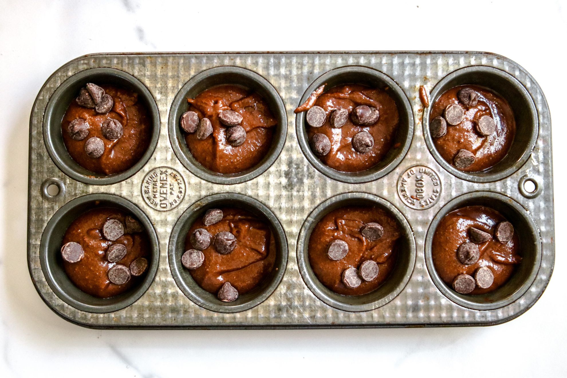 This is an overhead horizontal image of a muffin tin with eight cavities filled with chocolate batter. Each chocolate batter is topped with about four chocolate chips. The cupcake pan sits on a white marble surface.