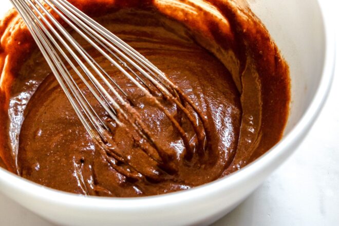 This is a horizontal image from a 3/4 view looking into a white bowl with chocolate batter. A whisk is in the bowl. The bowl sits on a white surface.