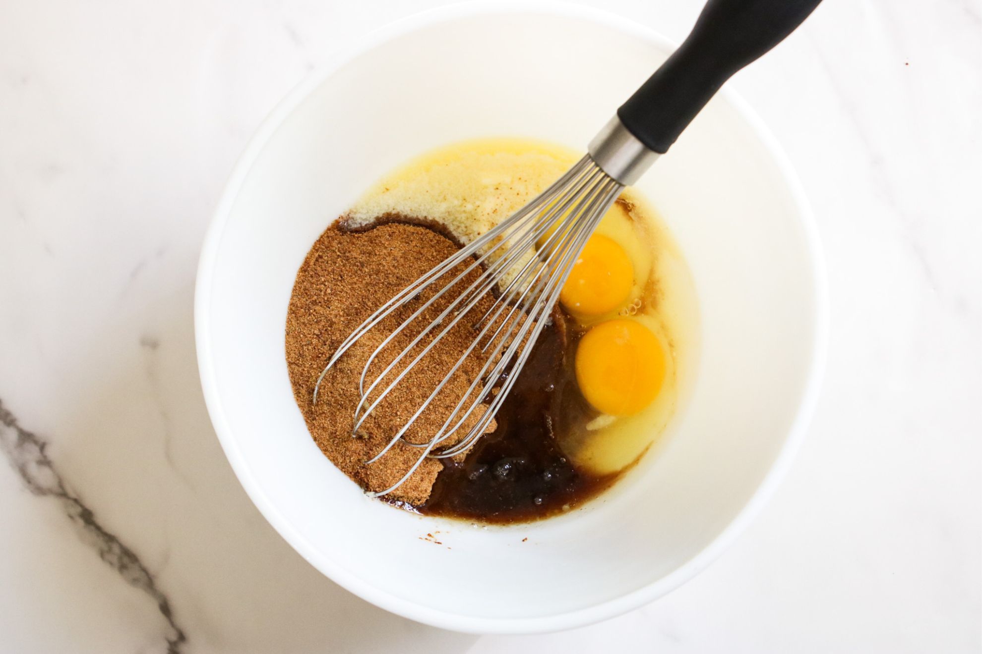 This is an overhead horizontal image of a white bowl with butter, eggs, coconut sugar, and vanilla extract in it. The bowl sits on a white marble surface. A whisk is in the bowl, leaning against the side with the handle pointing toward the top right of the image.