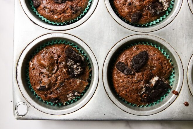 This is an overhead horizontal image of silver muffin tin on a white surface. Chocolate muffins have teal liners around them and have crushed oreos in each muffin cup.