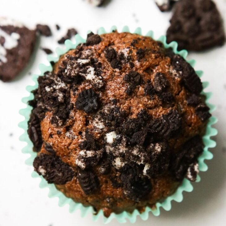 This is an overhead vertical image of a chocolate muffin with a teal muffin liner. The muffin is topped with crushed oreos. The muffin sits on a white surface with crushed oreos on the whit surface above the muffin.